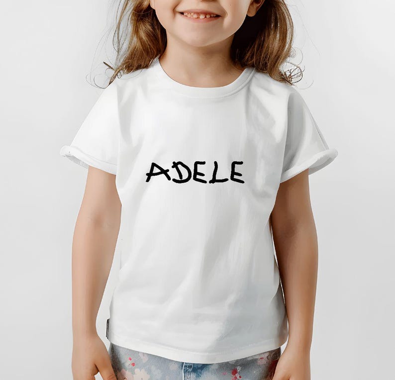 K&ouml;nnte beinhalten: Wei&szlig;es T-Shirt mit dem Namen "ADELE" in schwarzer, handschriftlicher Schrift. Das Shirt hat kurze &Auml;rmel und einen Rundhalsausschnitt. Das Model tr&auml;gt eine Hose mit Muster.