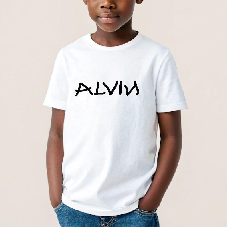 K&ouml;nnte beinhalten: Ein wei&szlig;es T-Shirt mit dem Namen "ALVIN" in schwarzer, stilisierter Schrift. Das T-Shirt hat einen Rundhalsausschnitt und wird von einer Person getragen. Die Person tr&auml;gt eine blaue Jeans.