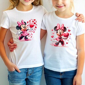 May include: Two white t-shirts featuring a Minnie Mouse graphic. The cartoon character is wearing a pink polka-dot dress and bow, holding a heart. Red hearts surround the character. The shirts are paired with blue jeans.