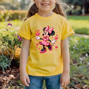 May include: A yellow t-shirt featuring a Minnie Mouse graphic. Minnie Mouse is depicted in a pink dress with a polka dot pattern, a matching bow, and holding a heart. The shirt is worn by a person wearing blue jeans.