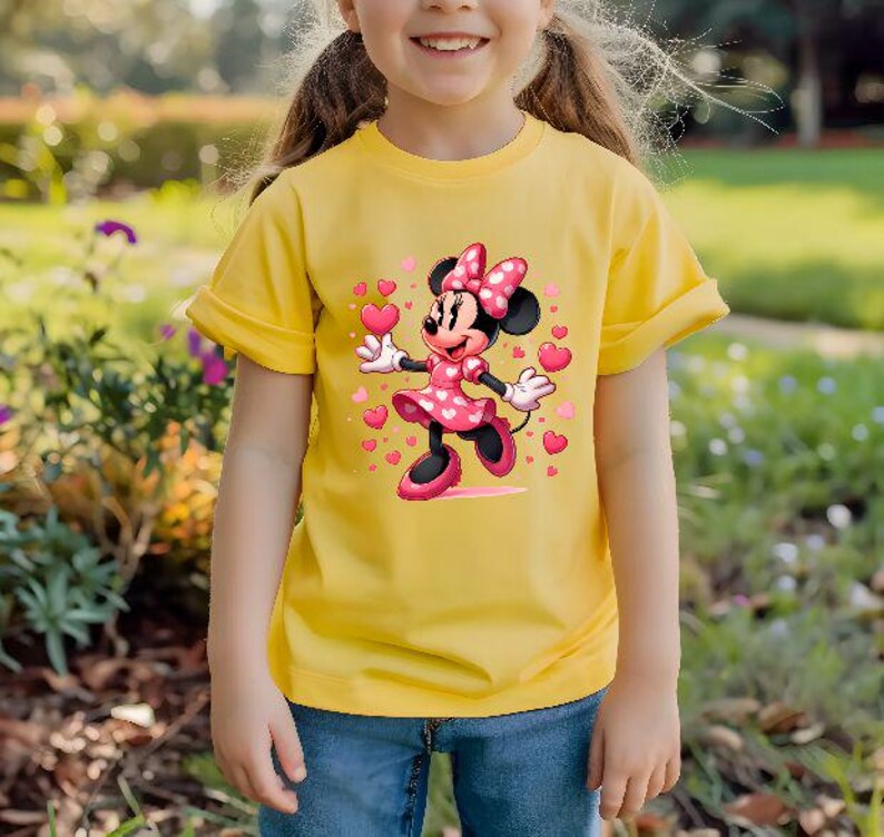 May include: A yellow t-shirt featuring a Minnie Mouse graphic. Minnie Mouse is depicted in a pink dress with a polka dot pattern, a matching bow, and holding a heart. The shirt is worn by a person wearing blue jeans.