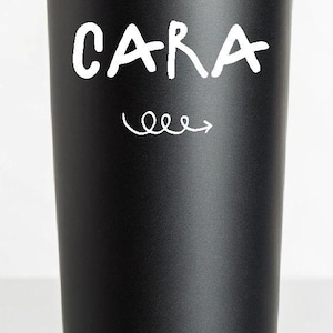 K&ouml;nnte beinhalten: Schwarzer isolierter Becher mit dem Namen "CARA" in wei&szlig;er Schrift, begleitet von einem wei&szlig;en Pfeil. Der Becher ist zylindrisch, hat einen schwarzen Deckel und eine matte Oberfl&auml;che. Wiederverwendbarer Trinkbeh&auml;lter.