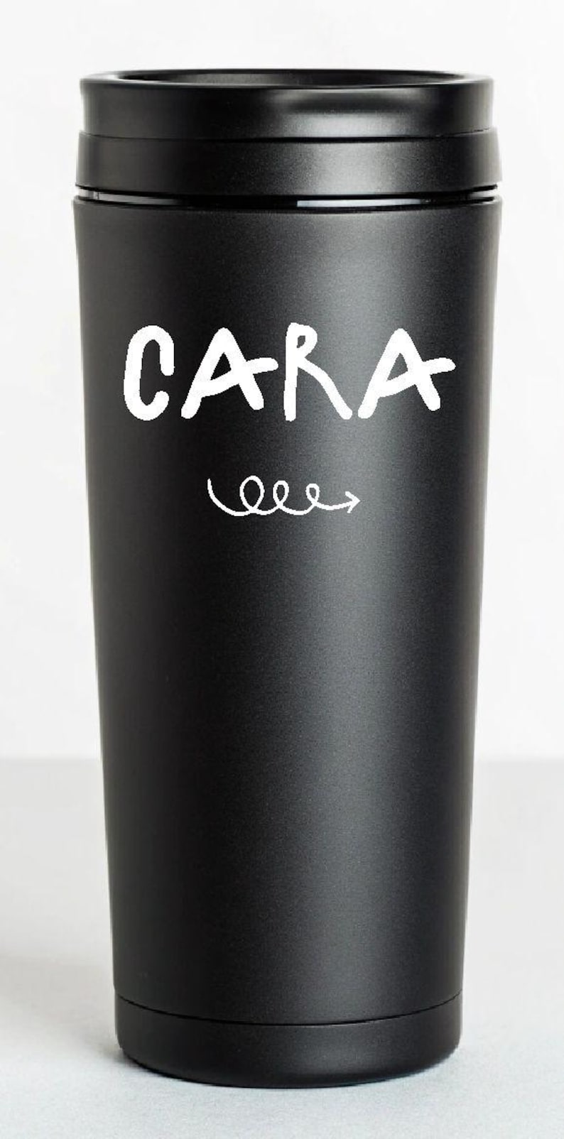 K&ouml;nnte beinhalten: Schwarzer isolierter Becher mit dem Namen "CARA" in wei&szlig;er Schrift, begleitet von einem wei&szlig;en Pfeil. Der Becher ist zylindrisch, hat einen schwarzen Deckel und eine matte Oberfl&auml;che. Wiederverwendbarer Trinkbeh&auml;lter.