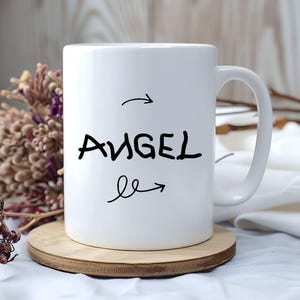K&ouml;nnte beinhalten: Wei&szlig;e Keramiktasse mit dem Wort "ANGEL" in schwarzer Handschrift, mit Pfeilen, die auf das Wort zeigen und davon weg. Die Tasse steht auf einem runden Holzuntersetzer. Getrocknete Blumen sind im Hintergrund.