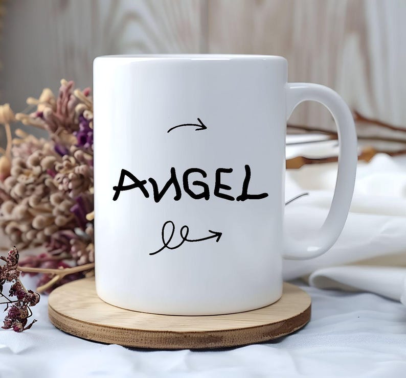 K&ouml;nnte beinhalten: Wei&szlig;e Keramiktasse mit dem Wort "ANGEL" in schwarzer Handschrift, mit Pfeilen, die auf das Wort zeigen und davon weg. Die Tasse steht auf einem runden Holzuntersetzer. Getrocknete Blumen sind im Hintergrund.