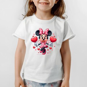 May include: White t-shirt featuring a cartoon Minnie Mouse design. Minnie Mouse is wearing a pink polka dot dress and bow, holding two red heart-shaped balloons. Small red hearts surround the character, creating a festive and cheerful look.