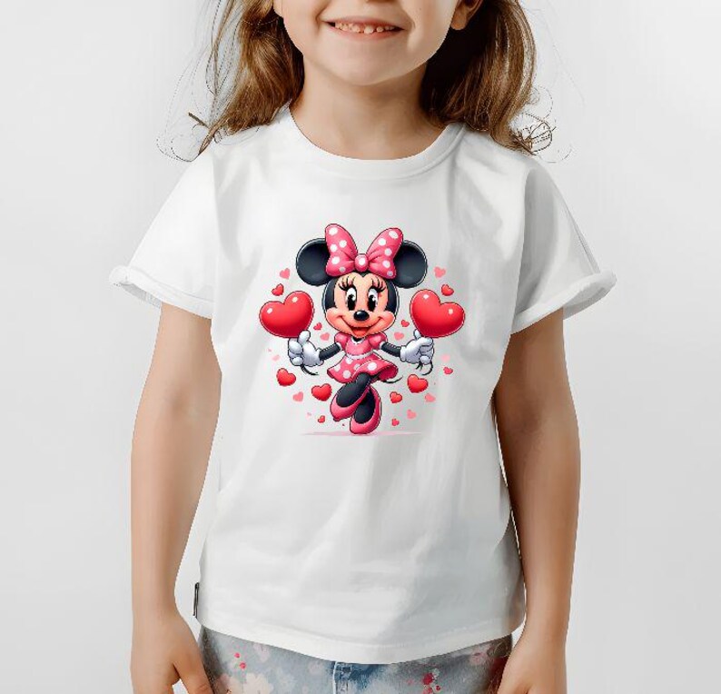 May include: White t-shirt featuring a cartoon Minnie Mouse design. Minnie Mouse is wearing a pink polka dot dress and bow, holding two red heart-shaped balloons. Small red hearts surround the character, creating a festive and cheerful look.