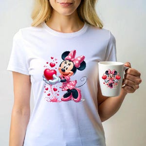 May include: White t-shirt featuring a Minnie Mouse graphic holding a red heart, with smaller hearts around her. A matching Minnie Mouse mug is held in the hand. The design includes pink polka dots and black accents.