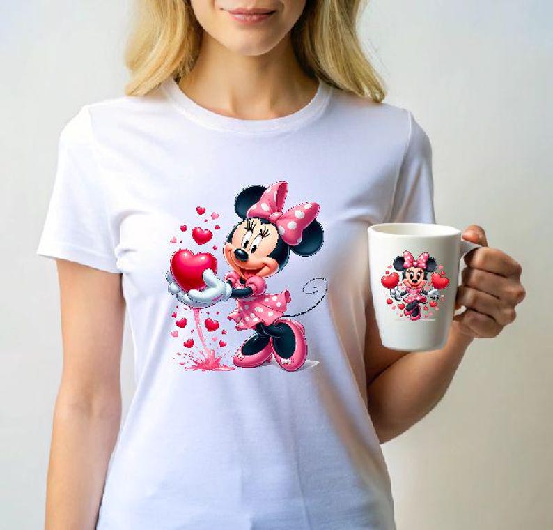 May include: White t-shirt featuring a Minnie Mouse graphic holding a red heart, with smaller hearts around her. A matching Minnie Mouse mug is held in the hand. The design includes pink polka dots and black accents.