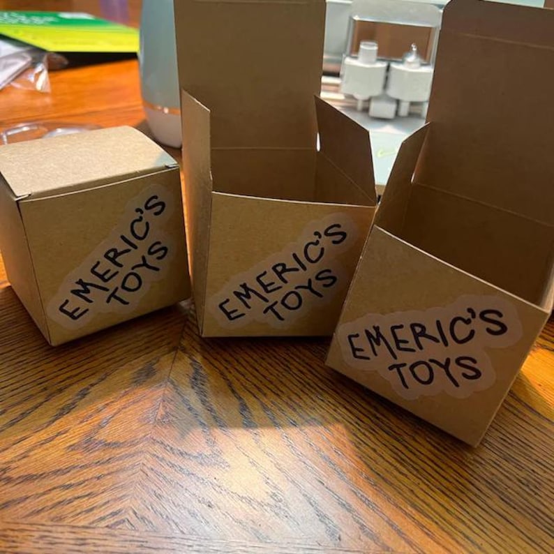 K&ouml;nnte beinhalten: Drei kleine, offene Pappschachteln mit dem Text "EMERIC'S TOYS" auf Aufklebern. Die Schachteln sind auf einer Holzoberfl&auml;che angeordnet. Eine Schachtel ist geschlossen, w&auml;hrend die anderen beiden offen sind und ihr Inneres zeigen.