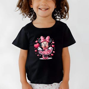 May include: Black t-shirt featuring a Minnie Mouse graphic. Minnie is wearing a pink polka-dot dress and holding a heart-shaped balloon. Pink hearts surround Minnie. The shirt has short sleeves and a round neckline.