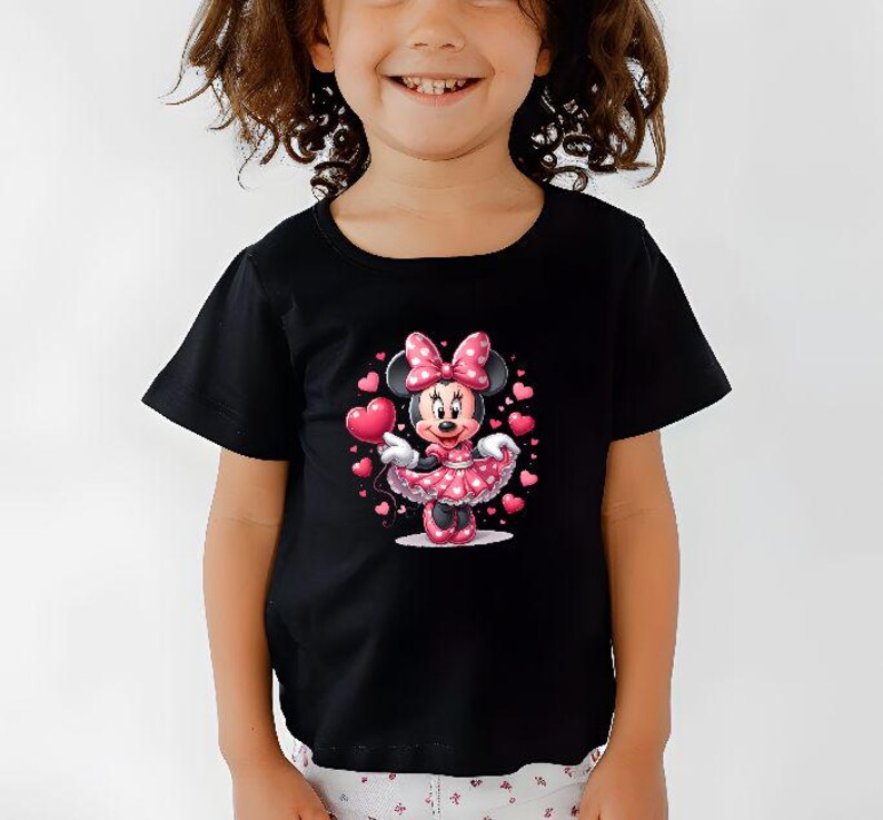 May include: Black t-shirt featuring a Minnie Mouse graphic. Minnie is wearing a pink polka-dot dress and holding a heart-shaped balloon. Pink hearts surround Minnie. The shirt has short sleeves and a round neckline.