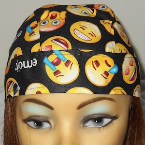 May include: A black bandana with a yellow and white emoji pattern. The bandana has a yellow emoji with a tear on the left side, a yellow emoji with a laughing mouth, and a yellow emoji with a heart-shaped eye. The bandana is worn on a mannequin's head.
