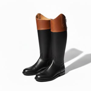 May include: A pair of knee-high riding boots featuring a smooth black lower section and a contrasting tan leather cuff at the top. The boots have a rounded toe, a low heel, and a vertical strap detail on the outer side of the tan collar.