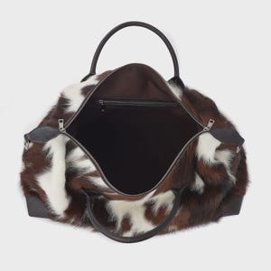 May include: Open duffel bag in brown and white cowhide, featuring black leather handles and trim. The bag has a zip closure and a roomy interior. Designed for travel, it has a classic, luxurious appearance.