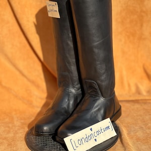 May include: A pair of black leather riding boots. The boots are tall, with a smooth finish and a rounded toe. The boots are resting on a dark, circular surface. The text on the tag reads "London Costume".