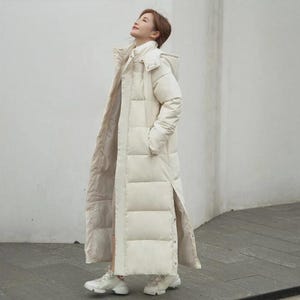 Può includere: Un lungo cappotto imbottito color crema con cappuccio. Il cappotto arriva alle caviglie e presenta un collo alto e uno spacco verticale sul lato. Il cappotto è indossato con scarpe da ginnastica bianche.