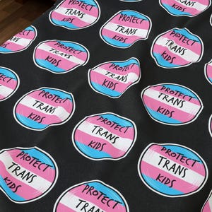 May include: Black blanket with repeating circular designs. Each design features the text "PROTECT TRANS KIDS" in a white, pink, and blue color scheme. The blanket is on a bed with a white pillow.