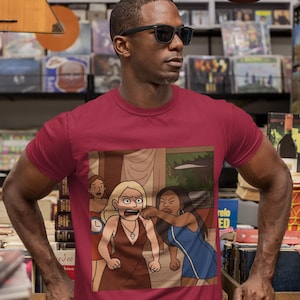 May include: A maroon short-sleeved t-shirt featuring a graphic illustration of three cartoon characters in a dramatic scene. One character with blonde hair wears a brown dress and a cross necklace, while another in a blue dress appears to be pulling her hair.