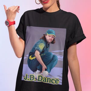 May include: A black short-sleeved t-shirt featuring a graphic print of a person crouching while wearing a green and yellow sports uniform and a matching cap. The text J.D. Dance is printed in yellow and blue lettering at the bottom of the graphic.