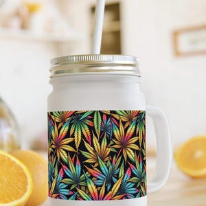 Op de afbeelding: Een drinkglas in de vorm van een weckpot van matglas met een zilverkleurig metalen deksel en een wit rietje. De buitenkant is voorzien van een herhalend patroon van veelkleurige cannabisbladeren in geel, roze, blauw en groen op een zwarte achtergrond.