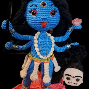 Puede incluir: Muñeca de ganchillo azul que representa a la diosa hindú Kali, con múltiples brazos, una corona y un collar. La muñeca tiene pelo negro, ojos rojos y una lengua roja. Se encuentra sobre una flor de loto rosa y sostiene un peine y una flor de loto.