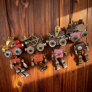 May include: A collection of steampunk-style pendants and charms, featuring miniature train and robot designs. The pendants are crafted from wood and metal, with intricate details like gears, wheels, and decorative accents in gold, silver, and red. They hang against a wooden background.