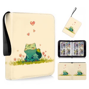 May include: A cream-coloured trading card binder featuring a cartoon green creature sitting in grass with small flowers and floating hearts. The binder includes a black wrist strap, a zippered closure, and internal plastic sleeves for cards.