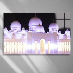May include: A photograph of a white mosque at night, illuminated with purple and gold lights. The mosque features multiple domes and minarets, with arched entryways and intricate architectural details. The image is a close-up shot, capturing the details of the building.