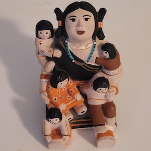 May include: A ceramic figurine depicting a woman with black hair in pigtails, adorned with a turquoise necklace, holding and surrounded by several small children. The children wear outfits in shades of orange, brown, and white. The figurine has a cream-colored base.