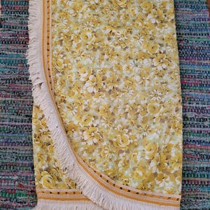 May include: A round, floral-patterned beach towel with a yellow and white flower design. The towel has a cream-colored fringe and a yellow and brown border. The towel is folded in half, showing the floral pattern and fringe.