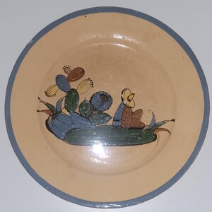 May include: A round ceramic plate with a light tan base and a dark gray rim. The center features a hand-painted illustration of a person wearing a yellow sombrero sitting next to a green cactus with blue, yellow, and brown fruit accents.