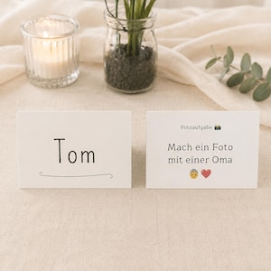 Könnte beinhalten: Zwei weiße Karten mit schwarzem Text auf einer beigen Oberfläche. Eine Karte zeigt "Tom" mit einer Linie darunter. Die andere Karte zeigt "Fotoaufgabe" und "Mach ein Foto mit einer Oma" mit einem Kamera-Emoji, einer Person und einem Herz.