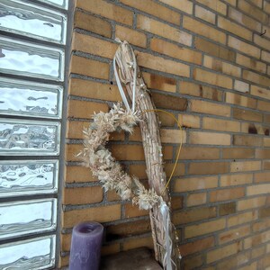 May include: A heart-shaped wreath made of dried flowers and foliage, with a wooden base and a yellow wire accent. A decorative piece with a rustic aesthetic, it is displayed near a brick wall and a purple candle.