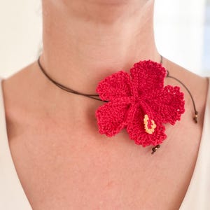 Op de afbeelding: Een ketting met een felrode gehaakte hibiscusbloem. De bloem, met gele accenten, is bevestigd aan een bruin koord. Deze handgemaakte ketting is een opvallend accessoire, perfect voor een tropische uitstraling.