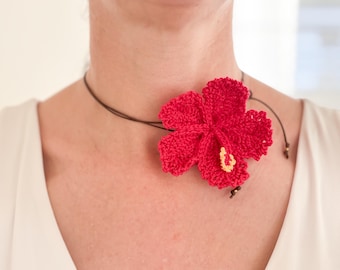 May include: A vibrant red crochet hibiscus flower necklace. The flower, with yellow accents, is attached to a brown cord. The necklace is a handmade accessory, perfect for adding a touch of tropical flair to any outfit.
