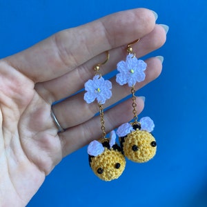 May include: A pair of gold dangle earrings featuring crocheted white flowers and yellow and black bumblebees.