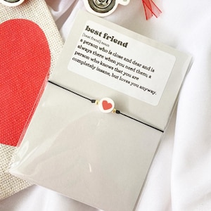 May include: A white card with the definition of "best friend" printed on it. The card is decorated with a red heart charm bracelet.