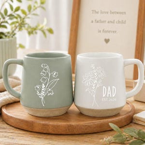 Könnte beinhalten: Zwei Keramikbecher mit floralen Designs. Einer ist salbeigrün mit einer weißen Blumenillustration, der andere ist weiß mit einem Blumendesign und dem Wort "DAD" und "EST. 2026". Beide Becher haben einen beigefarbenen Boden und einen gebogenen Griff.