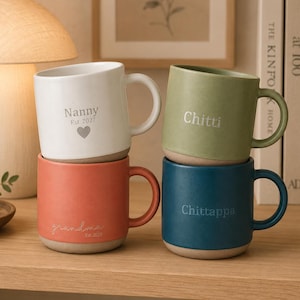 Könnte beinhalten: Vier Keramikbecher mit matter Oberfläche in Weiß, Salbeigrün, Terrakotta und Dunkelblau. Der weiße Becher trägt den Text Nanny Est. 2027 und ein Herz. Der terrakottafarbene Becher zeigt grandma Est. 2027. Der grüne Becher trägt Chitti und der blaue Chittappa.