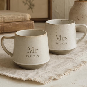 Könnte beinhalten: Zwei cremefarbene Keramikbecher mit Henkeln. Einer trägt die Aufschrift "Mr EST. 2026" und der andere "Mrs EST. 2026". Die Becher haben eine gesprenkelte Oberfläche und einen grauen Sockel. Sie stehen auf einem beigen Leinentuch.