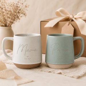 Könnte beinhalten: Zwei Keramikbecher, einer cremefarben und einer salbeigrün, mit den Worten "Mama" und "Papa" in Schreibschrift. Jeder Becher hat einen braunen Boden und einen Henkel. Die Becher stehen neben einer Geschenkbox mit einer Schleife.