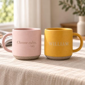 Peut inclure: Un mug rose avec le texte Choose calm. - Lily et un mug jaune avec le texte WILLIAM. Les deux mugs présentent une finition mate mouchetée, une anse arrondie et une base grise contrastante. Ils sont placés côte à côte sur une surface texturée.