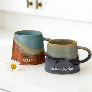 Könnte beinhalten: Zwei Keramikbecher mit einem einzigartigen Farbverlauf. Ein Becher ist türkis und orange mit dem Aufdruck "BELLA". Der andere Becher ist grau und blau mit dem Aufdruck "Mother's Day Gift". Vor den Bechern liegt eine weiße Gänseblümchen.