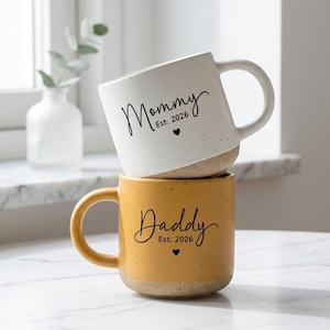 Puede incluir: Dos tazas de cerámica apiladas. La taza superior es blanca con la palabra "Mommy" y "Est. 2026" impresas en negro, con un pequeño corazón. La taza inferior es amarilla con la palabra "Daddy" y "Est. 2026" impresas en negro, con un pequeño corazón.