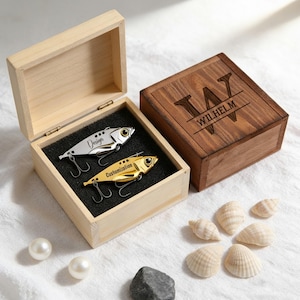 Könnte beinhalten: Zwei Angelköder, einer silberfarben und einer goldfarben, in einer Holzkiste mit schwarzem Schaumstoff. Der silberne Köder trägt das Wort "Design" und der goldene Köder das Wort "Customization". Eine zweite Holzkiste ist mit dem Namen "WILHELM" graviert.