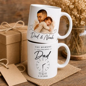 Könnte beinhalten: Zwei weiße Keramikbecher übereinander gestapelt. Der obere Becher zeigt eine Illustration von Vater und Kind mit dem Text "Dad & Noah". Der untere Becher hat ein Zifferblatt-Design und den Text "THE MOMENT you became Dad" mit Datum und Uhrzeit.