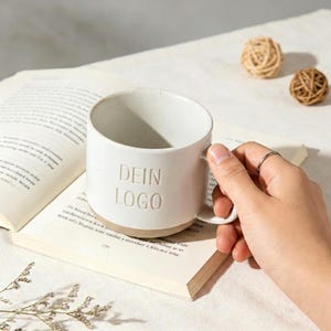 Könnte beinhalten: Eine weiße Keramik-Tasse mit dem Text "DEIN LOGO" auf der Seite. Die Tasse steht auf einem offenen Buch. Die Tasse hat einen hellbraunen Boden und einen Henkel. Zwei dekorative Kugeln sind im Hintergrund.