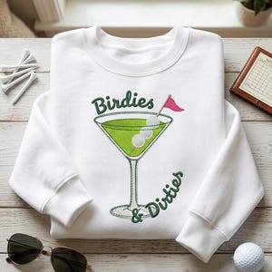 Puede incluir: Una sudadera blanca de manga larga con un bordado que muestra una copa de martini llena de líquido verde. Dentro de la copa hay tres pelotas de golf blancas en un palillo con una pequeña bandera rosa. El texto dice Birdies & Dirties en verde.