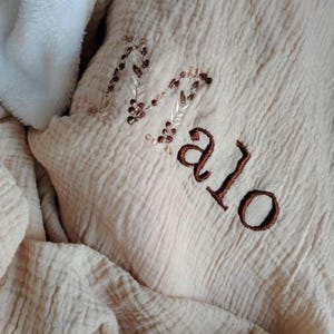 Peut inclure: Une couverture beige texturée avec le nom "Malo" brodé en fil marron. La couverture a une bordure blanche douce et moelleuse. La broderie comprend des paillettes et un motif de feuilles, ajoutant une touche décorative à la couverture pour bébé.